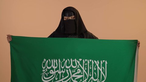 Showing loyalty to the country, our beginning day February 22, Saudi National Day September 23, wearing traditional dress on a national occasion, Flag Day March 11, close-up portrait of a Saudi Arabian Gulf woman wearing a burqa and abaya holding the Kingdom's flag and looking at the camera with gestures of pride, floral background