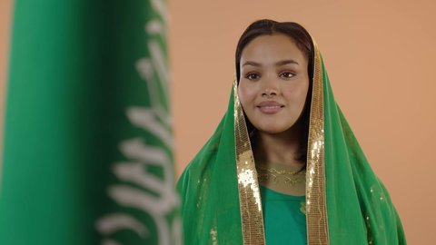 Our beginning day is February 22, celebrating Saudi National Day on September 23, Flag Day on March 11, wearing the traditional costume of the central region on a national occasion, close-up portrait of a Saudi Arabian Gulf woman wearing a green shayla and bisht standing in front of the Kingdom's flag, floral background