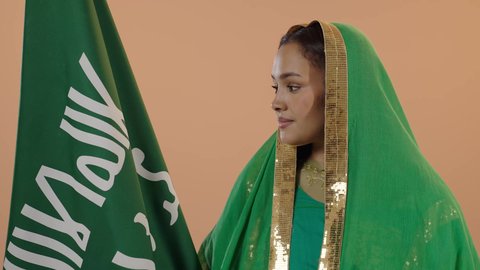 Celebrating the day of our beginning in 1727 AD, standing upright and raising the founding flag with expressions of pride and boasting, gestures of joy and happiness, the anniversary of the founding of the first Saudi state, February 22, a close-up portrait of a Saudi Arabian Gulf woman wearing a sheila and bisht, a floral background