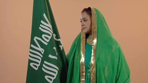 Looking at the flag with pride, celebrating the day we began in 1727 AD, standing up straight and raising the founding flag with expressions of pride and boasting, gestures of joy and happiness, the anniversary of the founding of the first Saudi state, February 22, a close-up portrait of a Saudi Arabian Gulf woman wearing a sheila and a bisht, a floral background