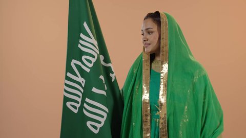 Celebrating the day of our beginning in 1727 AD, standing upright and raising the founding flag with expressions of pride and boasting, gestures of joy and happiness, the anniversary of the founding of the first Saudi state, February 22, a close-up portrait of a Saudi Arabian Gulf woman wearing a sheila and bisht, a floral background