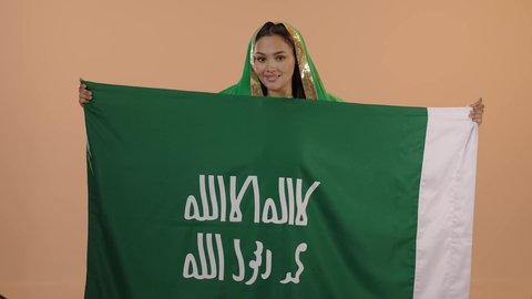 النظر إلى الكاميرا بفخر وسرور، الاحتفال بيوم بدينا ١٧٢٧م، الوقوف باستقامة ورفع علم التأسيس بتعابير الشموخ والتباهي، ايماءات البهجة والسرور، ذكرى تأسيس الدولة السعودية الاولى ٢٢ فبراير، بورتريه لامرأة عربية خليجية سعودية ترتدي الشيلة والبشت الاخضر، خلفية زهرية