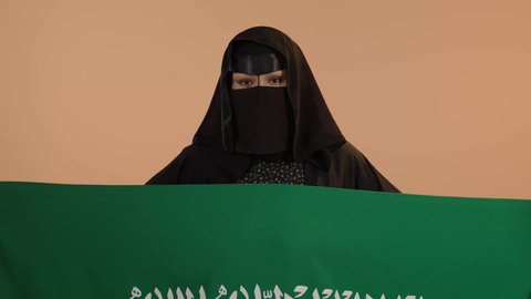 Showing loyalty to the country, our beginning day February 22, Saudi National Day September 23, wearing traditional dress on a national occasion, Flag Day March 11, close-up portrait of a Saudi Arabian Gulf woman wearing a burqa and abaya holding the Kingdom's flag and looking at the camera with gestures of pride, floral background