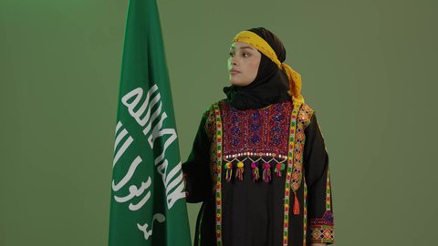 يوم بدينا 1727 م، ذكرى تأسيس الدولة السعودية الأولى 22 فبراير، ارتداء أزياء المنطقة الوسطى، بورتريه لإمرأة عربية خليجية سعودية مبتسمة ترتدي الكرتة والهامة تنظر إلى علم يوم التأسيس بفخر، الاحتفال بالمناسبات والأعياد الوطنية الشعبية، خلفية خضراء، كروما