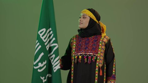 يوم بدينا 1727 م، ذكرى تأسيس الدولة السعودية الأولى 22 فبراير، ارتداء أزياء المنطقة الوسطى، بورتريه لإمرأة عربية خليجية سعودية مبتسمة ترتدي الكرتة والهامة تنظر إلى علم يوم التأسيس بفخر، الاحتفال بالمناسبات والأعياد الوطنية الشعبية، خلفية خضراء، كروما