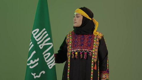 حب الوطن والانتماء إليه، يوم بدينا 1727 م، ذكرى تأسيس الدولة السعودية الأولى 22 فبراير، ارتداء أزياء المنطقة الوسطى، بورتريه لإمرأة عربية خليجية سعودية مبتسمة ترتدي الكرتة والهامة تنظر إلى علم يوم التأسيس بفخر، المناسبات والأعياد الوطنية الشعبية، خلفية خضراء، كروما