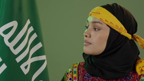 المناسبات والأعياد الوطنية الشعبية، حب الوطن والانتماء إليه، يوم بدينا 1727 م، ذكرى تأسيس الدولة السعودية الأولى 22 فبراير، ارتداء أزياء المنطقة الوسطى، بورتريه مقرب لإمرأة عربية خليجية سعودية ترتدي الكرتة والهامة تنظر إلى علم يوم التأسيس بفخر، خلفية خضراء، كروما