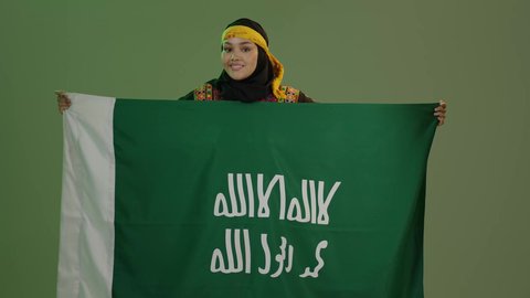 ذكرى تأسيس الدولة السعودية الأولى 22 فبراير، يوم بدينا 1727 م، ارتداء أزياء المنطقة الوسطى، بورتريه لإمرأة عربية خليجية سعودية مبتسمة ترتدي الكرتة والهامة تحمل بيدها علم يوم التأسيس، الاحتفال بالمناسبات والأعياد الوطنية الشعبية، خلفية خضراء، كروما