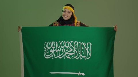 اليوم الوطني السعودي 23 سبتمبر, النظر إلى الكاميرا بفخر، الاعتزاز بالتراث التاريخي، ارتداء زي المنطقة الوسطى، الاحتفال بالمناسبات الوطنية، يوم العلم 11 مارس، بورتريه لإمرأة عربية خليجية سعودية مبتسمة ترتدي الكرتة والهامة تحمل علم المملكة, خلفية خضراء، كروما