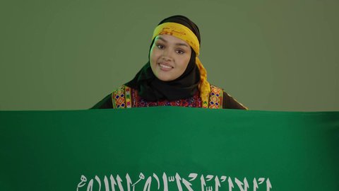 Celebrating national occasions, Saudi National Day September 23, looking at the camera with pride, taking pride in historical heritage, wearing the Central Region costume, Flag Day March 11, close-up portrait of a smiling Saudi Arabian Gulf woman wearing a kurta and a hat holding the Kingdom's flag, green background, chroma key