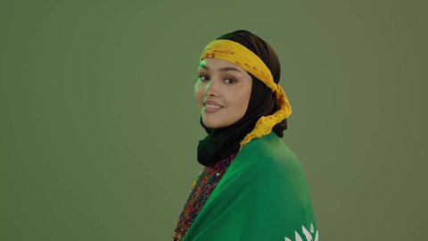 ذكرى اليوم الوطني السعودي 23 سبتمبر, النظر إلى الكاميرا بفخر، الاعتزاز بالتراث التاريخي، ارتداء زي المنطقة الوسطى، يوم العلم 11 مارس، بورتريه مقرب لإمرأة عربية خليجية سعودية مبتسمة ترتدي الكرتة والهامة تضع علم المملكة على كتفيها, خلفية خضراء، كروما