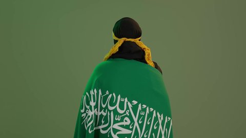 الاحتفال بذكرى اليوم الوطني السعودي 23 سبتمبر, يوم العلم 11 مارس، الاعتزاز بالتراث التاريخي، ارتداء زي المنطقة الوسطى، بورتريه من الخلف لإمرأة عربية خليجية سعودية مبتسمة ترتدي الكرتة والهامة تضع علم المملكة على كتفيها, خلفية خضراء، كروما