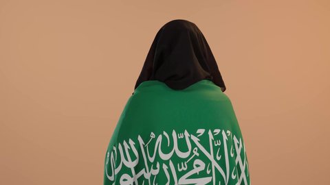 الفخر والاعتزاز بالانتماء للبلاد، يوم بدينا22 فبراير, اليوم الوطني السعودي 23 سبتمبر، ارتداء الزي الشعبي في مناسبة وطنية، يوم العلم 11 مارس، بورتريه من الخلف لامرأة عربية خليجية سعودية ترتدي البرقع والعباءة تضع على كتفيهاعلم المملكة، خلفية زهرية