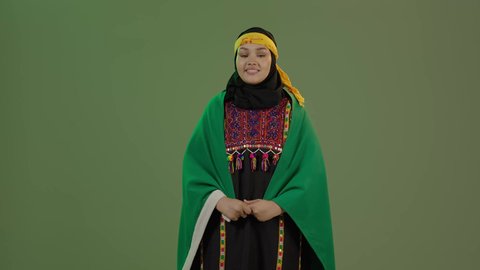 يوم العلم 11 مارس، ذكرى اليوم الوطني السعودي 23 سبتمبر, النظر إلى الكاميرا بفخر، الاعتزاز بالتراث التاريخي، ارتداء زي المنطقة الوسطى، بورتريه لإمرأة عربية خليجية سعودية مبتسمة ترتدي الكرتة والهامة تضع علم المملكة على كتفيها, خلفية خضراء، كروما