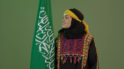 النظر إلى العلم بفخر، الاعتزاز بالتراث التاريخي، ارتداء زي المنطقة الوسطى، الاحتفال بالمناسبات الوطنية، اليوم الوطني السعودي 23 سبتمبر, يوم العلم 11 مارس، بورتريه لإمرأة عربية خليجية سعودية مبتسمة ترتدي الكرتة والهامة تحمل علم المملكة, خلفية خضراء، كروما