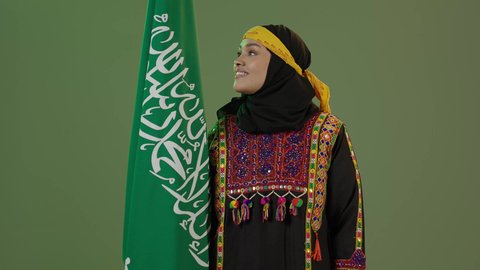 Saudi National Day September 23, looking at the flag with pride, taking pride in historical heritage, wearing the Central Region costume, celebrating national occasions, Flag Day March 11, portrait of a smiling Saudi Arabian Gulf woman wearing a kurta and a hat holding the Kingdom's flag, green background, chroma key