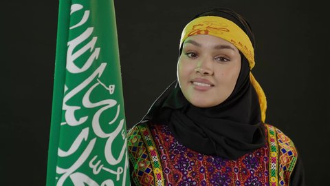 Looking at the camera with pleasure, celebrating national occasions, Saudi National Day September 23, the day we started 1727 AD, pride in historical heritage, Flag Day March 11, wearing the Central Region costume, close-up portrait of a Saudi Arabian Gulf woman wearing a kurta and a hat holding the Kingdom's flag, black background