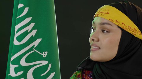 ارتداء زي المنطقة الوسطى، النظر إلى العلم بفخر، الاحتفال بالمناسبات الوطنية، اليوم الوطني السعودي 23 سبتمبر, يوم بدينا 1727م, الفخر بالتراث التاريخي، يوم العلم 11 مارس، بورتريه مقرب لإمرأة عربية خليجية سعودية ترتدي الكرتة والهامة تحمل علم المملكة, خلفية سوداء