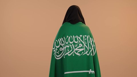 Pride and pride in belonging to the country, the day we started February 22, Saudi National Day September 23, wearing traditional dress on a national occasion, Flag Day March 11, a portrait from the back of a Saudi Arabian Gulf woman wearing a burqa and abaya, placing the flag of the Kingdom on her shoulders, a floral background
