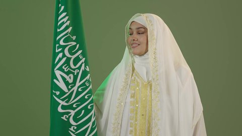 السعادة بالانتماء للهوية السعودية، الأزياء الشعبية التقليدية، اليوم الوطني السعودي 23 سبتمبر، يوم العلم 11 مارس، بورتريه مقرب لامرأة عربية خليجية سعودية مبتسمة ترتدي الشيلة والنشل تمسك علم المملكة بإيماءات الفخر، ارتداء زي المنطقة الشرقية، خلفية خضراء، كروما