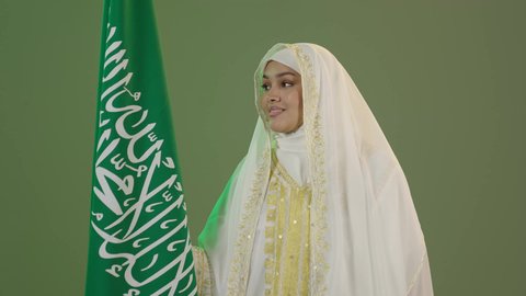 Traditional folk costumes, Saudi National Day September 23, looking at the camera with an expression of pride and loftiness, Flag Day March 11, close-up portrait of a smiling Saudi Arabian Gulf woman wearing a sheyla and a neshel holding the Kingdom's flag, wearing the Eastern Province costume, green background, chroma key