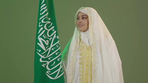 السعادة بالانتماء للهوية السعودية، الأزياء الشعبية التقليدية، اليوم الوطني السعودي 23 سبتمبر، يوم العلم 11 مارس، بورتريه مقرب لامرأة عربية خليجية سعودية مبتسمة ترتدي الشيلة والنشل تمسك علم المملكة بإيماءات الفخر، ارتداء زي المنطقة الشرقية، خلفية خضراء، كروما