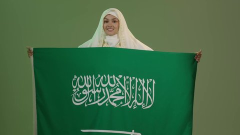 شعار الهوية الوطنية وراية الإسلام والتوحيد، الأزياء الشعبية التقليدية، اليوم الوطني السعودي 23 سبتمبر، يوم العلم 11 مارس، بورتريه لامرأة عربية خليجية سعودية مبتسمة ترتدي الشيلة والنشل تمسك علم المملكة بإيماءات الفخر، ارتداء زي المنطقة الشرقية، خلفية خضراء، كروما