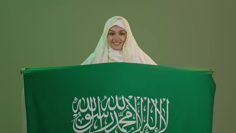 شعار الهوية الوطنية وراية الإسلام والتوحيد، الأزياء الشعبية التقليدية، اليوم الوطني السعودي 23 سبتمبر، يوم العلم 11 مارس، بورتريه لامرأة عربية خليجية سعودية مبتسمة ترتدي الشيلة والنشل تمسك علم المملكة بإيماءات الفخر، ارتداء زي المنطقة الشرقية، خلفية خضراء، كروما