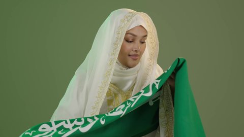 التباهي بإنجازات البلاد، النظر الى الكاميرا بتعابير الفخر، اليوم الوطني السعودي 23 سبتمبر، يوم العلم 11 مارس، بورتريه مقرب لامرأة عربية خليجية سعودية مبتسمة ترتدي الشيلة والنشل تقوم بتقبيل علم المملكة، ارتداء أزياء المنطقة الشرقية، خلفية خضراء، كروما