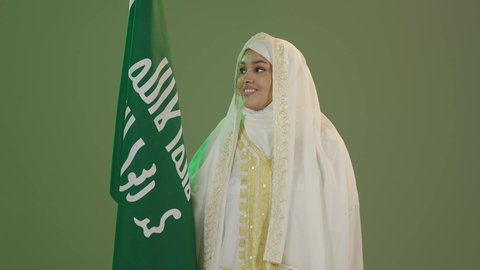 Celebrating national holidays, looking at the camera with expressions of pride and honor, wearing Eastern Province costumes, our day, February 22, portrait of a smiling Saudi Arabian Gulf woman wearing a sheila and a neshel holding the flag of the founding day of the first Saudi state 1727 AD, green background, chroma key
