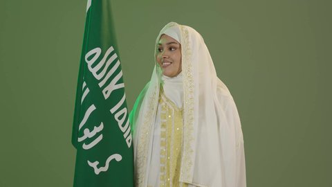 الاعتزاز بالانتماء للوطن، الاحتفال بالأعياد الوطنية، النظر إلى الكاميرا بتعابير السرور، ارتداء أزياء المنطقة الشرقية، يوم بدينا 22 فبراير، بورتريه لإمرأة عربية خليجية سعودية ترتدي الشيلة والنشل تحمل علم يوم تأسيس الدولة السعودية الأولى 1727 م، خلفية خضراء، كروما