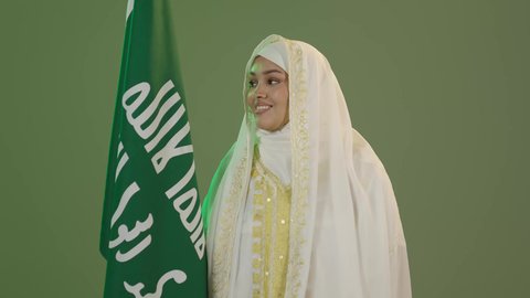 Pride in belonging to the homeland, celebrating national holidays, looking at the camera with expressions of pleasure, wearing the costumes of the Eastern Province, the day we started, February 22, a portrait of a Saudi Arabian Gulf woman wearing a sheila and a neshel carrying the flag of the founding day of the first Saudi state 1727 AD, green background, chroma key