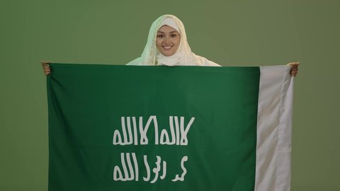 Looking at the camera with expressions of pride and honor, wearing the Eastern Province costumes, our day, February 22, celebrating national holidays, portrait of a smiling Saudi Arabian Gulf woman wearing a sheila and a neshel holding the flag of the founding day of the first Saudi state 1727 AD, green background, chroma key
