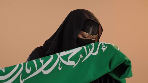 الاحتفال بذكرى اليوم الوطني السعودي 23 سبتمبر، النظر إلى الكاميرا بإيماءات الفخر، يوم العلم 11 مارس، يوم بدينا22 فبراير, ارتداء الزي الشعبي في مناسبة وطنية, بورتريه مقرب لامرأة عربية خليجية سعودية ترتدي البرقع والعباءة تقوم بتقبيل علم المملكة، خلفية وردية