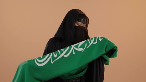 Raising the banner of monotheism and Islam, Flag Day March 11, Badina Day February 22, wearing traditional dress on a national occasion, celebrating Saudi National Day September 23, close-up portrait of a Saudi Arabian Gulf woman wearing a burqa and abaya hugging the Kingdom's flag, pink background