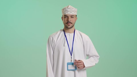 عرض البطاقة التعريفية ، الرجولة والاهتمام بالمظهر الخارجي, النظر إلى الكاميرا بتعابير البهجة والفرح، موك اب مفرغة باللون الابيض, بورتريه لرجل عربي خليجي عماني يرتدي الدشداشة و الكمة يقف بابتسامة, ارتداء بادج شخصي، خلفية خضراء