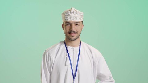 الرجولة والاهتمام بالمظهر الخارجي, عرض البطاقة التعريفية , النظر إلى الكاميرا بتعابير البهجة والفرح، موك اب مفرغة باللون الابيض, بورتريه مقرب لرجل عربي خليجي عماني يرتدي الدشداشة و الكمة يقف بابتسامة, ارتداء بادج شخصي، خلفية خضراء