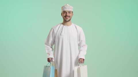 متعة التسوق فى سلطنة عمان, التبضع وأخذ الحاجات الضرورية, الاستفادة من الحسومات والتصفيات, أكياس الهدايا الورقية التجارية, بورتريه لشاب عربي خليجي عماني يرتدي الدشداشة و الكمة يمسك بيده حقائب المشتريات بايماءات بسعادة, خلفية خضراء