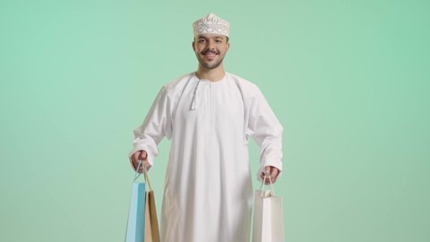التبضع وأخذ الحاجات الضرورية, الاستفادة من الحسومات والتصفيات, أكياس الهدايا الورقية التجارية, بورتريه لشاب عربي خليجي عماني يرتدي الدشداشة و الكمة يمسك بيده حقائب المشتريات بايماءات بسعادة, متعة التسوق فى المول, خلفية خضراء