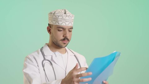 مراجعة نتيجة الفحص، بورتريه لطبيب عربي خليجي عماني يرتدي الدشداشة والكمة والزي الخاص بالمهنة يحمل ملف يقوم بقراءته يإيماءات التركيز والتمعن، العمل في القطاع الصحي في سلطنة عمان، إعداد التقرير الطبي، خلفية خضراء