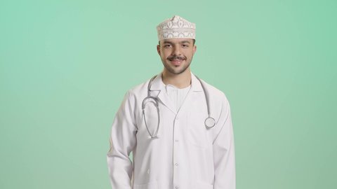 ايماءات القبول والموافقة, مفهوم الفحص الطبي، العمل في القطاع الصحي، تقديم الخدمات العلاجية للمرضى, بورتريه لطبيب عربي خليجي عماني يرتدي  المعطف الابيض و الكمة يضع السماعة الطبية يرفع يده بتعابير الاعجاب والرضى، خلفية خضراء
