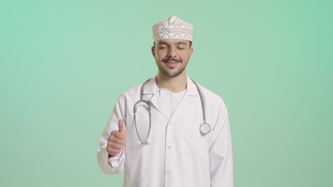 مفهوم الفحص الطبي، ايماءات القبول والموافقة، العمل في القطاع الصحي، تقديم الخدمات العلاجية للمرضى, بورتريه لطبيب عربي خليجي عماني يرتدي  المعطف الابيض و الكمة يضع السماعة الطبية يرفع يده بتعابير الاعجاب والرضى، خلفية خضراء