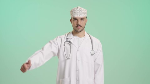 مفهوم التقييم السلبي والانزعاج، القلق والفشل في تحقيق أمر ما، بورتريه لطبيب عربي خليجي عماني يرتدي الكمه و المعطف الابيض و السماعات الطبية ينظر الى الكاميرا يشير بإصبع الابهام الى الاسفل بإيماءات الانزعاج، تعابير الرفض وعدم القبول والرضا،خلفية خضراء
