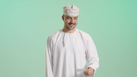 إمكانية تسديد الفواتير عبر بطاقة الصراف, الشراء والدفع عن طريق بطاقات الإئتمان البنكية, بورتريه لشاب عربي خليجي عماني يرتدي الدشداشة و الكمة يمسك بيده البطاقة الالكترونية ينظر الى الكاميرا بإيماءات السرور, خلفية خضراء