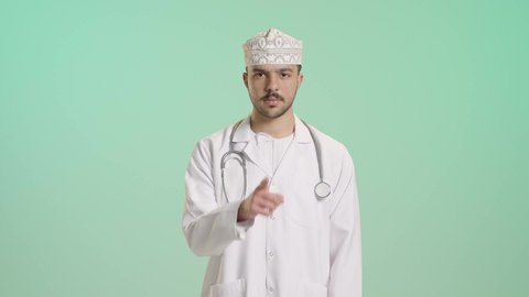 تعابير الجدية والحزم والغضب, الأمر بعدم الازعاج والسكوت والصمت، بورتريه لطبيب عربي خليجي عماني يرتدي الدشداشة و الكمة والمعطف الطبي يضع السماعة الطبية يعمل في القطاع الصحي يضع اصبع السبابة على فمه ينظر الى الكاميرا بإيماءات التوقف عن الكلام وطلب خفض الصوت، خلفية خضراء