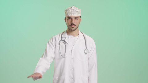 Raising the palm of the hand with expressions of stop and warning, a portrait of an Arab Gulf Omani doctor wearing a dishdasha, kumma, medical coat, and mask, holding a stethoscope, working in the health sector, with a green background.