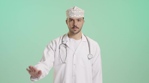 Raising the hand with gestures to stop talking and request silence, a portrait of an Arab Gulf Omani doctor wearing a dishdasha, kumma, medical coat, and mask, holding a stethoscope, working in the health sector, with a green background.