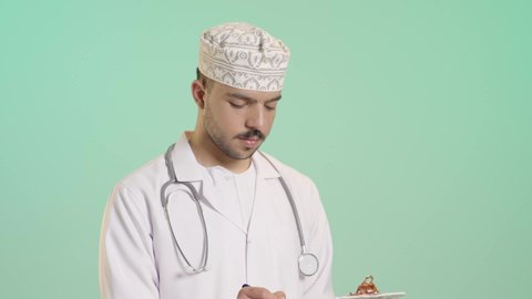 Recording test results in the medical record, gestures of memory and thought, a portrait of an Arab Gulf Omani doctor wearing a dishdasha, kumma, and white coat holding patient files in his hand, preparing medical reports, working in the health sector in the Sultanate of Oman, green background.
