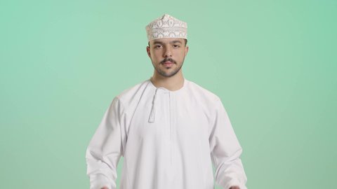 Gestures indicating satisfaction and approval, attention to elegance and appearance, a portrait of a young Arab Gulf Omani man wearing a dishdasha and kumma looking at the camera raising his index finger upwards with expressions of acceptance, joy, and happiness, green background.