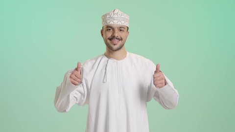 إيماءات الإعجاب والرضا، الاهتمام بالأناقة والمظهر الخارجي، بورتريه لشاب عربي خليجي عمانب يرتدي الدشداشة و الكمة ينظر الى الكاميرا يرفع إصبع السبابة للاعلى بتعابير القبول والفرح والسرور، خلفية خضراء